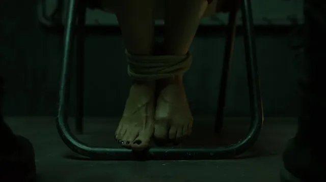 Úrsula Corberó shown with bare feet beside a chair in a dimly lit scene.