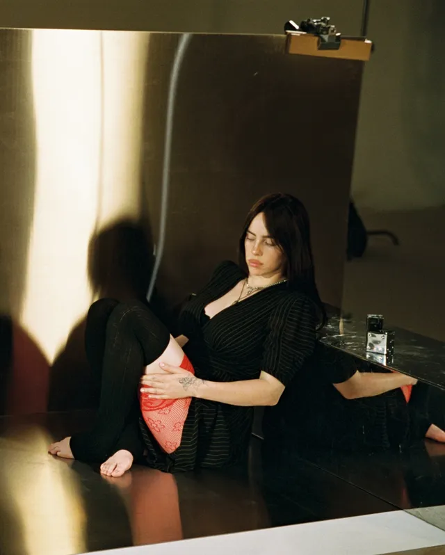 Billie Eilish a seated black outfit with a bare foot visible.