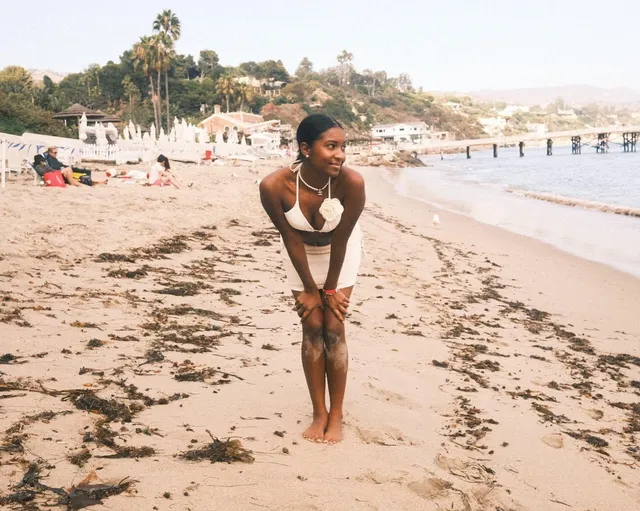 Carlacia Grant leaning forward barefoot on a beach in a white outfit.