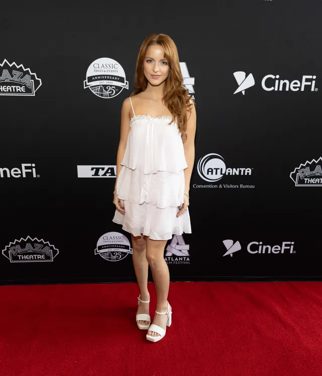 Mckenna Grace posing on an event carpet in a short white dress and heels.