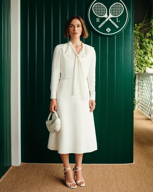 Keira Knightley posing beside a green doorway in a white dress and heels.