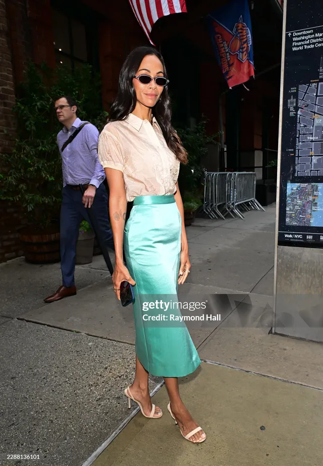 Zoe Saldaña standing outside in a white blouse and turquoise skirt.