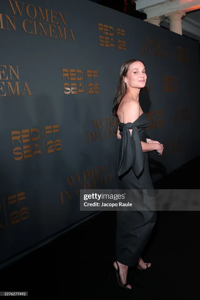 Alicia Vikander turning toward cameras in a black event dress.