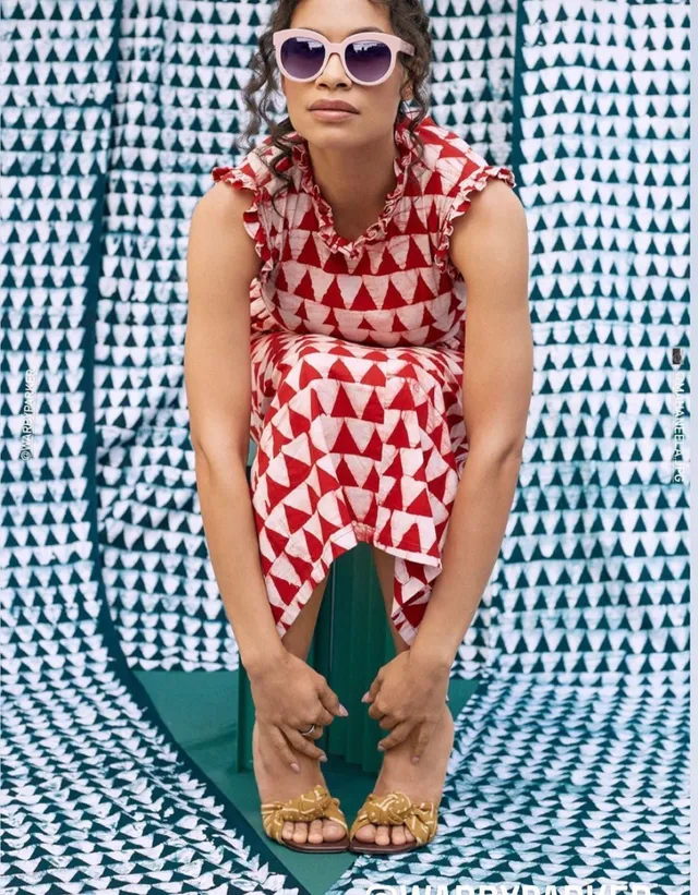 Rosario Dawson crouching in a red patterned dress and gold sandals.
