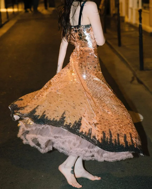 Dove Cameron posing barefoot in a patterned dress outdoors.