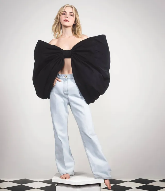 Kiernan Shipka standing barefoot on a scale in a large bow top and jeans.