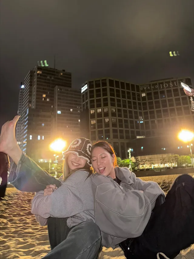 Lisa (Lalisa Manobal) sitting beside a companion outdoors at night during a fireworks display.