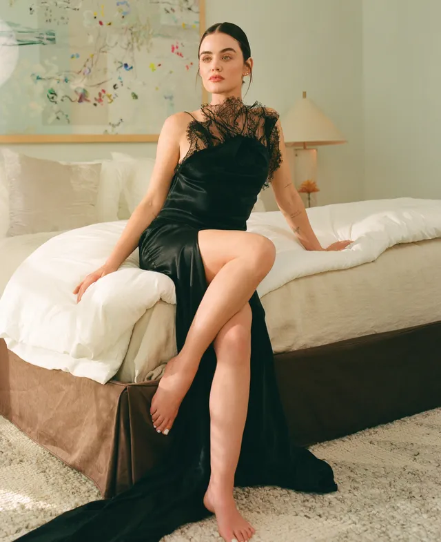 Lucy Hale sitting on a bed in a black feather-trimmed outfit.
