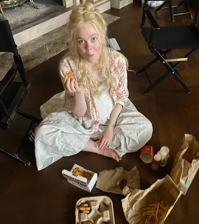 Elle Fanning sitting barefoot indoors in a pale outfit.