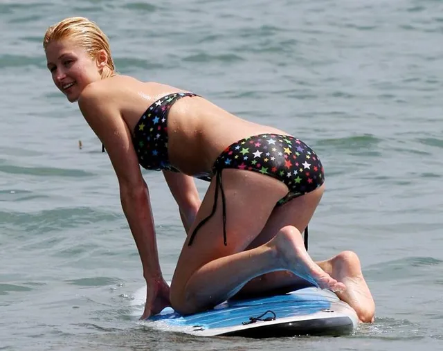 Paris Hilton kneeling barefoot on a surfboard.