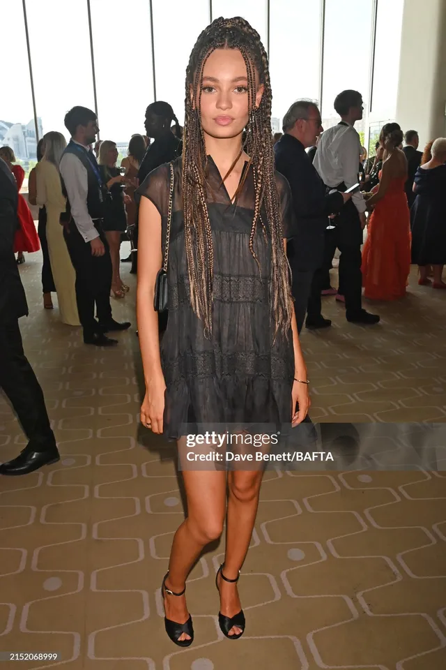 Nico Parker posing in a grey dress and black heels at an indoor event.
