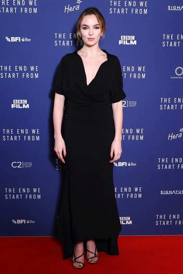 Jodie Comer posing against a blue premiere backdrop in a black gown.