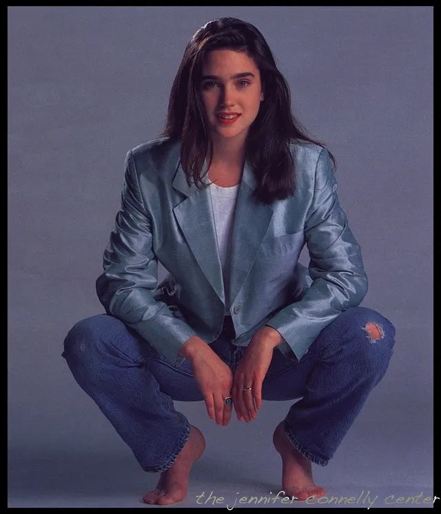 Jennifer Connelly crouching for a studio portrait in a jacket and jeans.