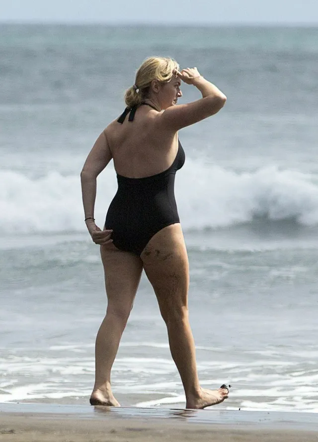 Kate Winslet walking barefoot on a beach in a black swimsuit.