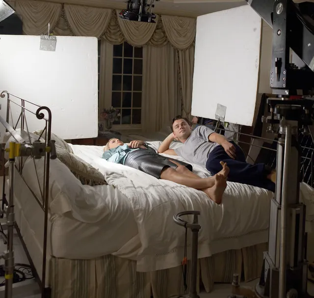 Margot Robbie reclining on a bed in a pale outfit beside another adult during a photo shoot.