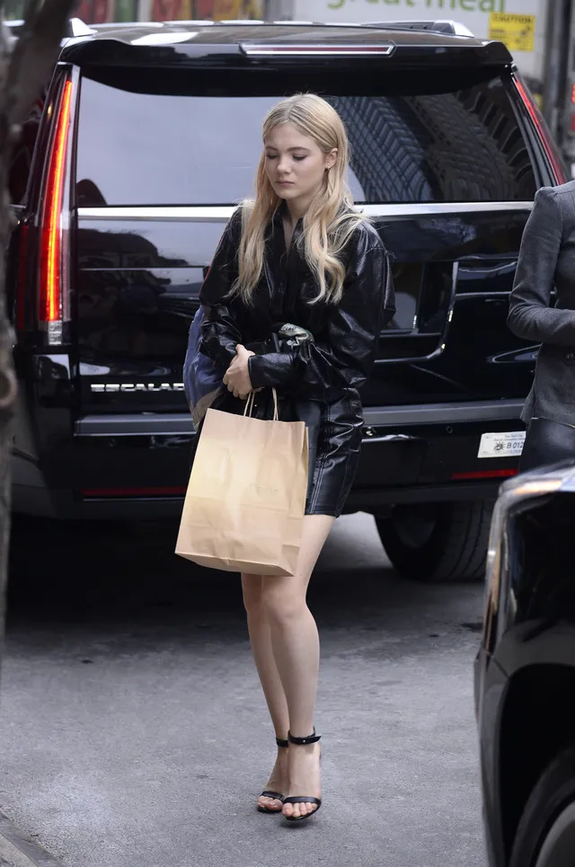 Freya Allan standing beside a vehicle in a black outfit and heels.