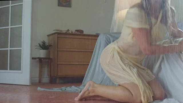Hilary Duff sitting barefoot beside a bed in a pale dress.