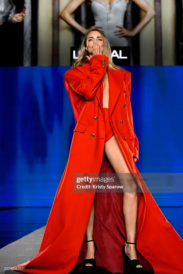 Cara Delevingne blowing a kiss at a fashion event in a red coat.