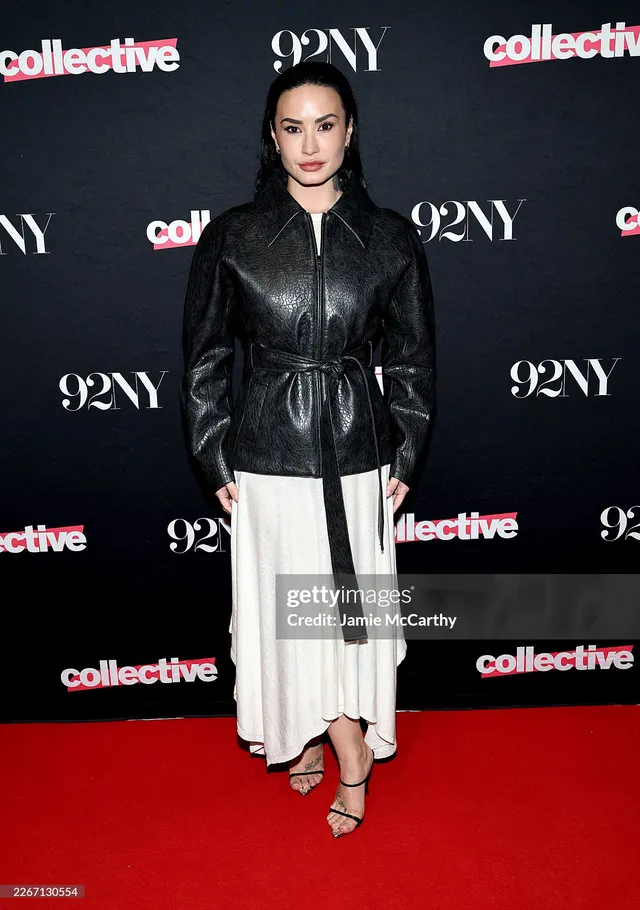 Demi Lovato standing at an event in a leather jacket, white skirt and heels.