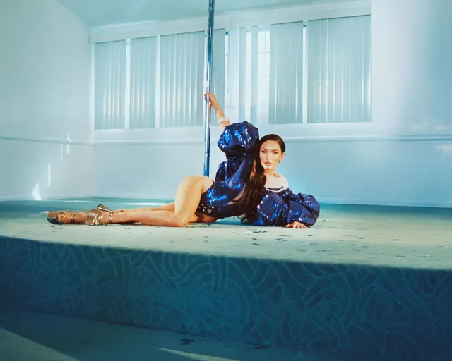 Megan Fox reclining barefoot beside an indoor pool wearing a blue outfit.
