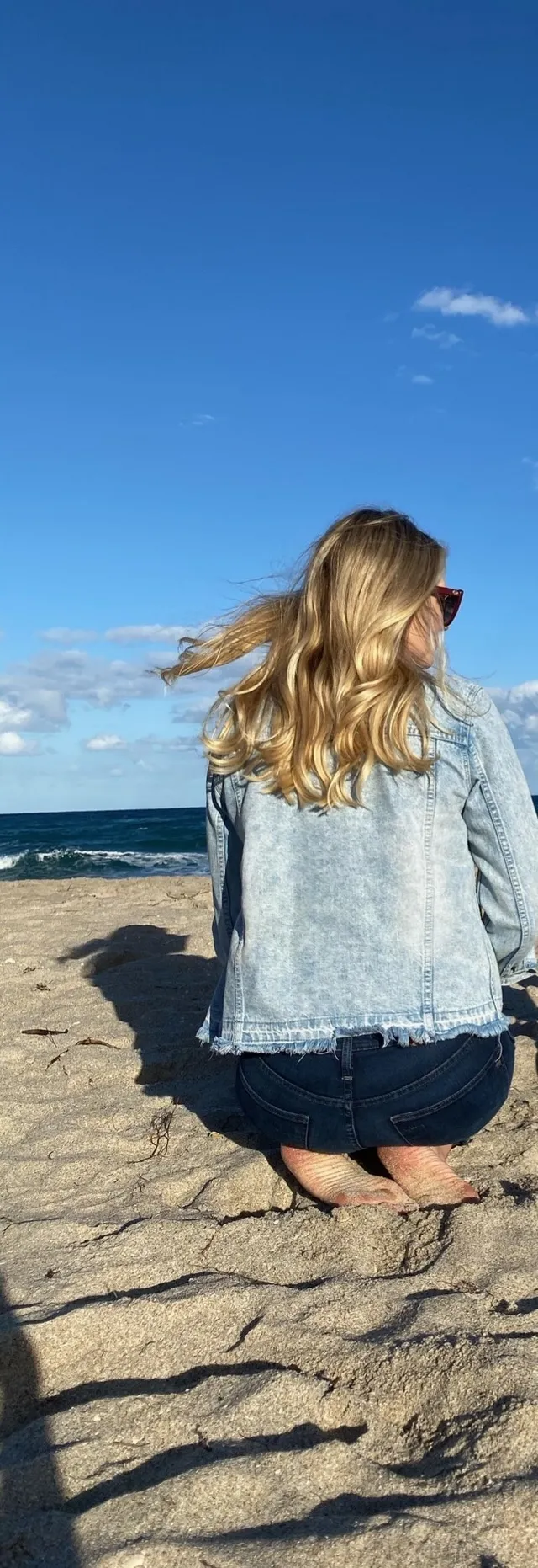 Kate Upton crouching barefoot on a sandy beach in rear view.
