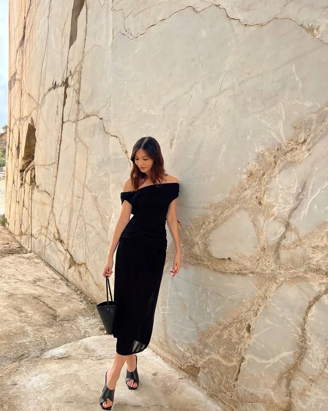 Gemma Chan standing beside a stone wall in a black dress.