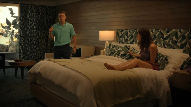 Alexandra Daddario sitting barefoot on a bed beside another person.