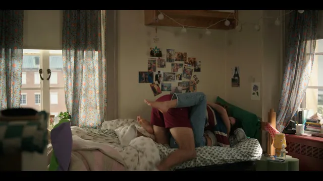 Lola Tung sitting barefoot on a bed beside a man.