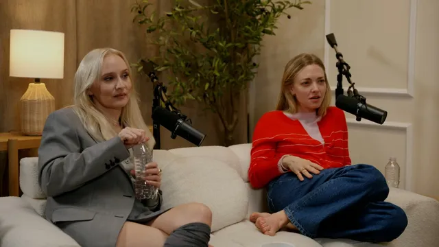 Amanda Seyfried sitting barefoot in jeans beside another guest on a couch.
