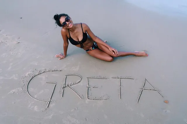 Greta Onieogou sitting barefoot on a beach beside a name drawn in the sand.
