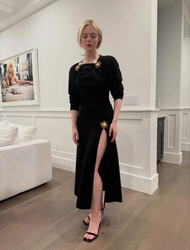 Elle Fanning posing in a long black dress and heeled sandals indoors.
