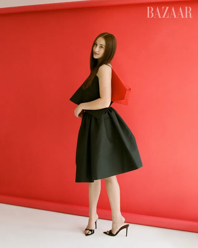 Lola Tung posing in a black dress and heels against a red studio backdrop.