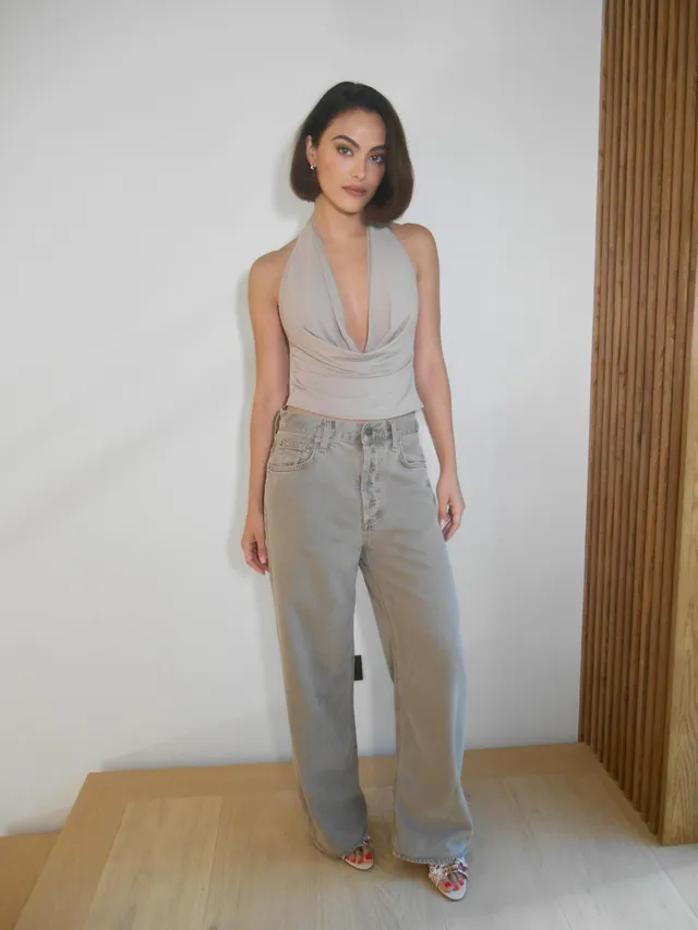 Camila Mendes wearing metallic heels and wide-leg gray trousers indoors.