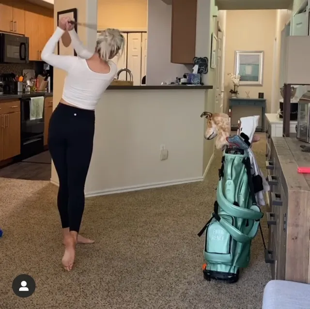 Paige Spiranac practising a golf swing barefoot on an indoor carpet.