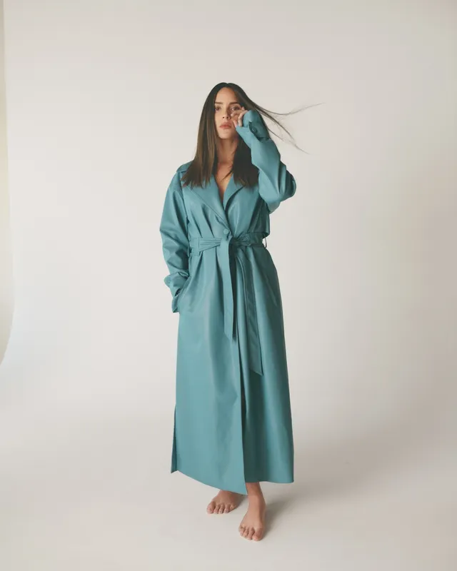 Adria Arjona standing barefoot in a long teal coat for a studio portrait.