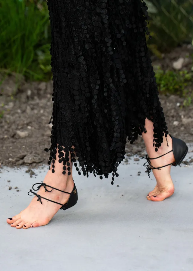 Margaret Qualley a close detail of black open toe heels beneath a fringed dress.