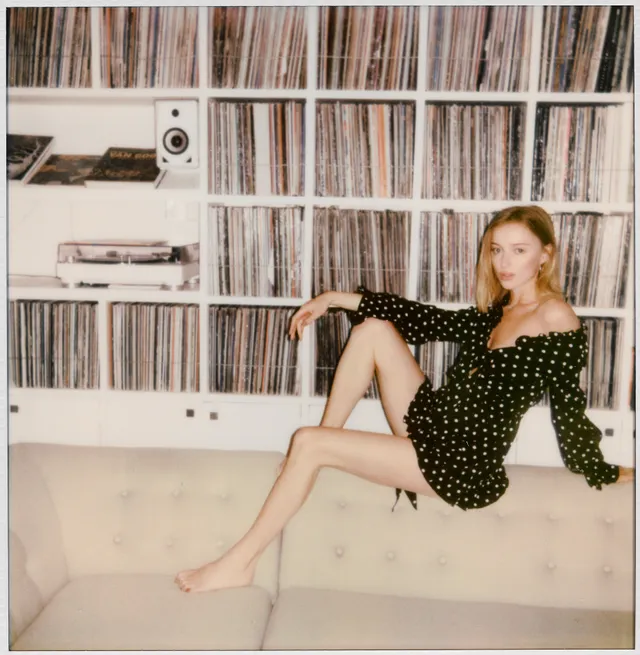 Phoebe Dynevor seated beside a wall of records in a polka-dot dress.