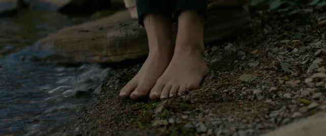 Anna Kendrick standing barefoot beside a shallow woodland stream.