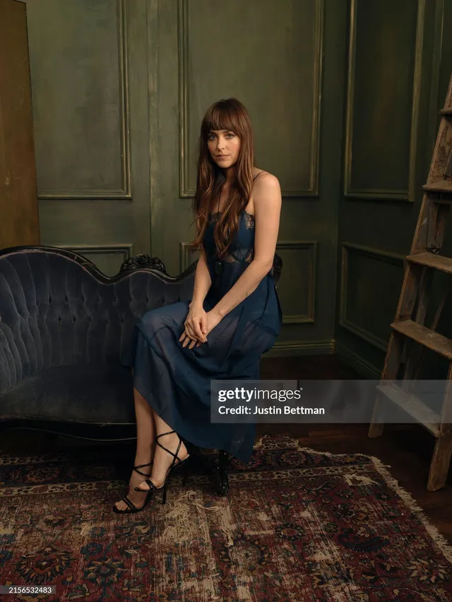 Dakota Johnson sitting on a sofa in a dark dress and strappy heels.