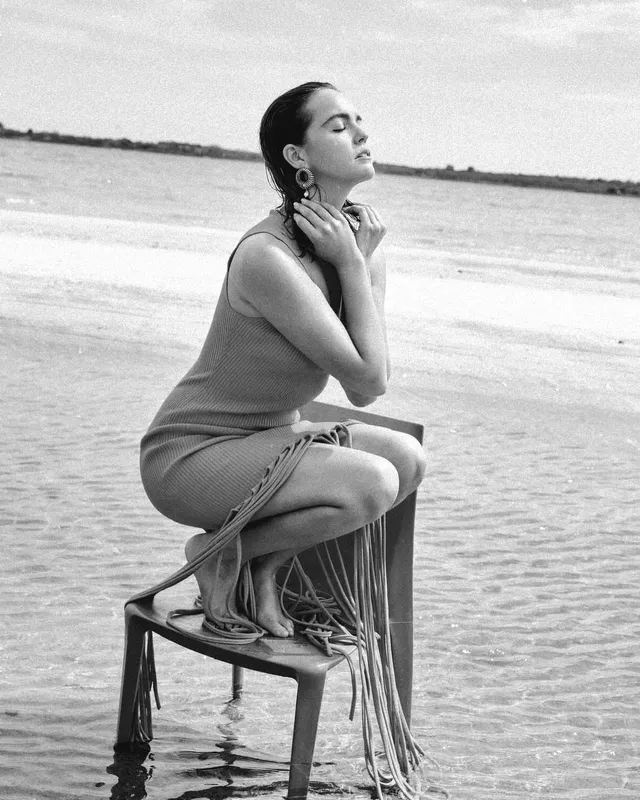 Bailee Madison sitting barefoot on a chair beside the water.