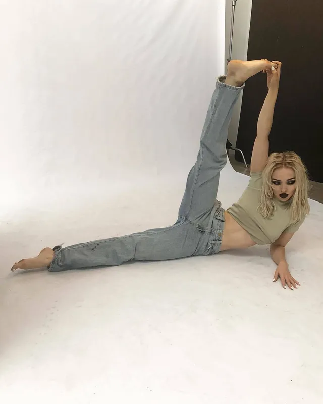 Dove Cameron stretching barefoot on a white studio floor.