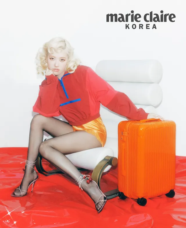Rosé sitting beside an orange suitcase in a red top, shorts and patterned high heels.