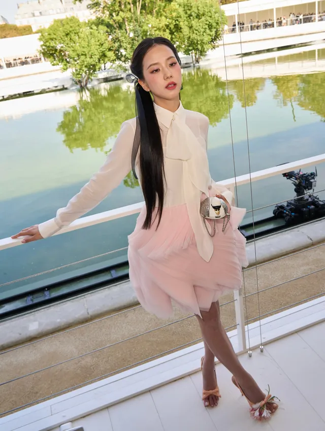 Jisoo standing on a balcony in pale heels, a white blouse and a pink skirt.